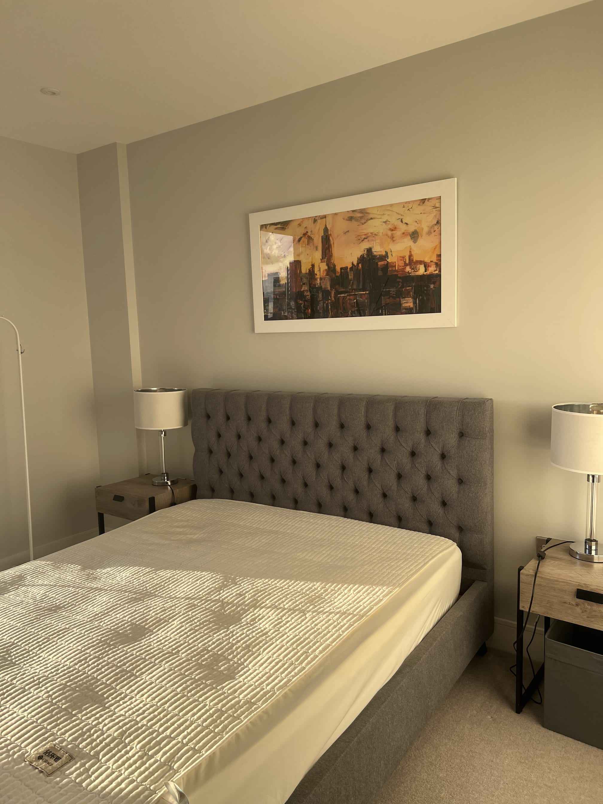 Made-up double bed with a grey buttoned headboard, bedside lamps and a city painting above