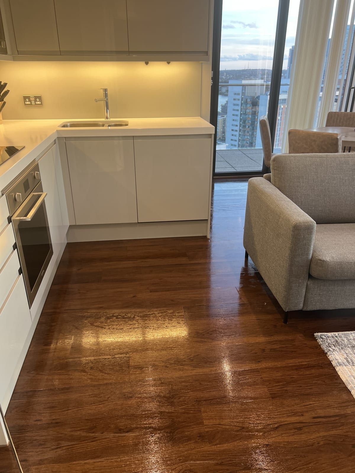 Open-plan flat with a cream kitchen, glossy wooden floor and a city view through tall windows
